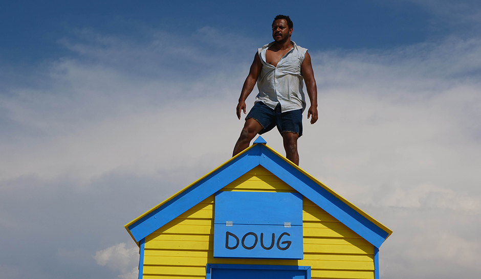 A man standing on top of a yellow and blue wooden house. The house has a nameplate reading "DOUG".
