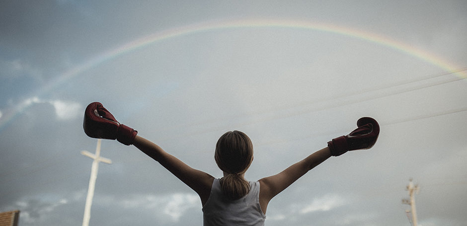 Girl wearing boxing gloves with arms thrown wide looking up to the sky. There is a rainbow above her and a white cross in the distance.