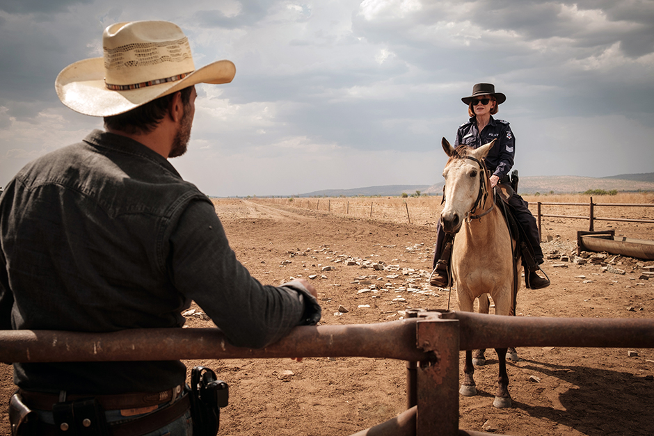 Judy Davis riding a horse with a casual coolness towards Aaron Pedersen with storm clouds and desert in the background
