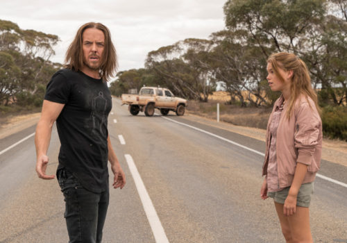 A man and girl standing on a bush road looking tired and exasperated with a haphazardly parked pickup truck in the background.