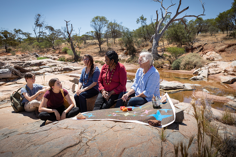 Yamaji Arts artist Wendy Jackamarra and her sister Glenda Jackamarra talk about Wendy's painting 'The Jewellery Box' to Lucia Richardson, Max Winton & Charmaine Green in the bush