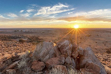 Outback with red earth, scrubland and rocks with sun hanging low over the horizon in a clear blue sky.