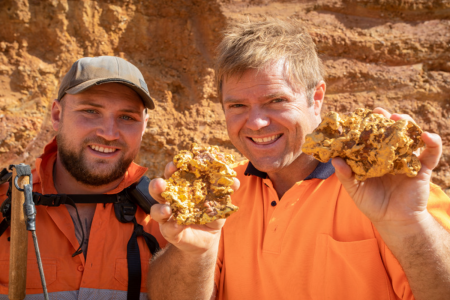 Two people in a dig site wearing high-vis clothing holding up gold nuggets